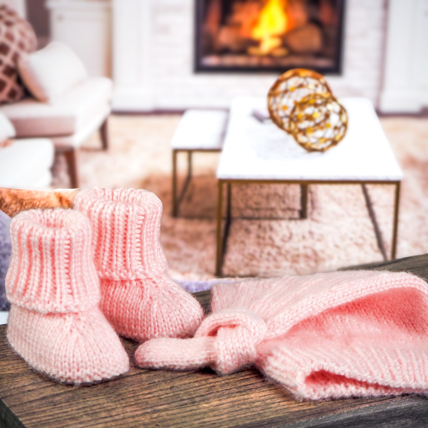 Pink knitted baby booties and a matching knotted baby hat, displayed on a wooden surface in front of a cozy living room setting with a fireplace and modern decor.