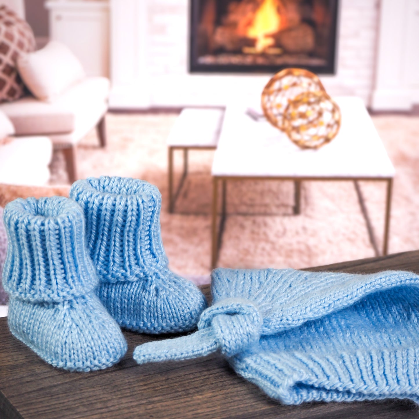 Light blue knitted baby booties with a matching knotted baby hat, displayed on a wooden surface with a warm living room background featuring a glowing fireplace and modern decor.