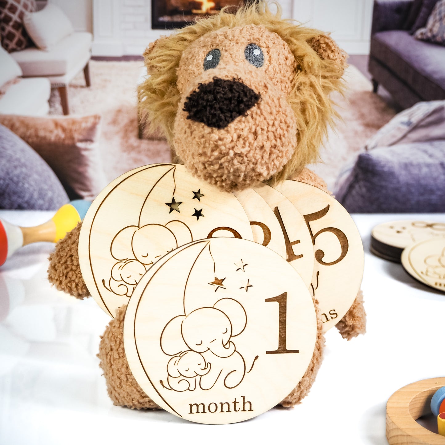 A plush lion toy holding a set of wooden baby milestone cards engraved with a sleeping elephant design, including "1 month" and other milestones, displayed in a cozy living room setting.