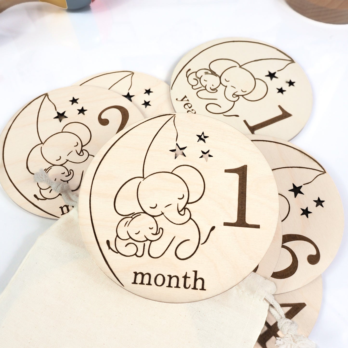 A collection of wooden baby milestone cards engraved with a sleeping elephant design, featuring "1 month" and other milestones, arranged on a white surface with a drawstring storage bag.