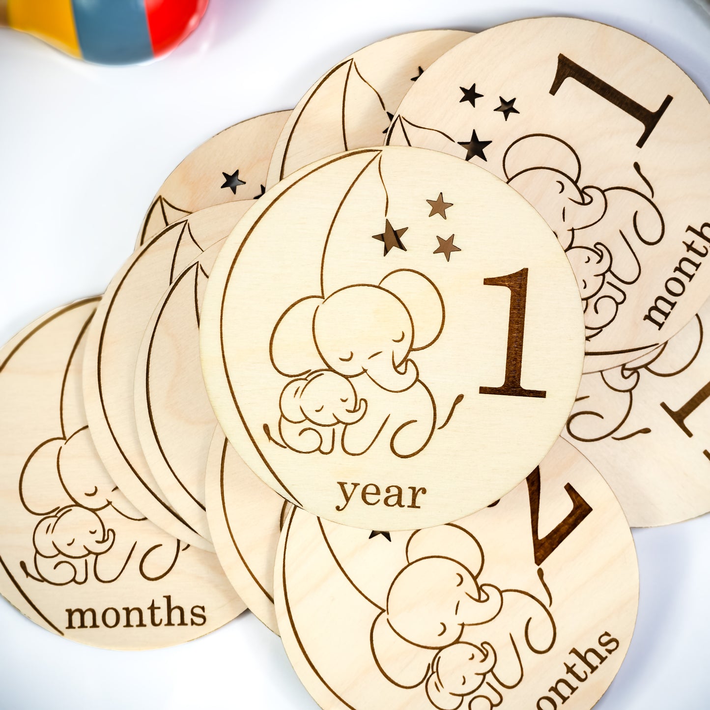 A stack of engraved wooden baby milestone cards with a sleeping elephant design, featuring milestones such as "1 year" and "months," displayed on a white surface with colorful background elements.