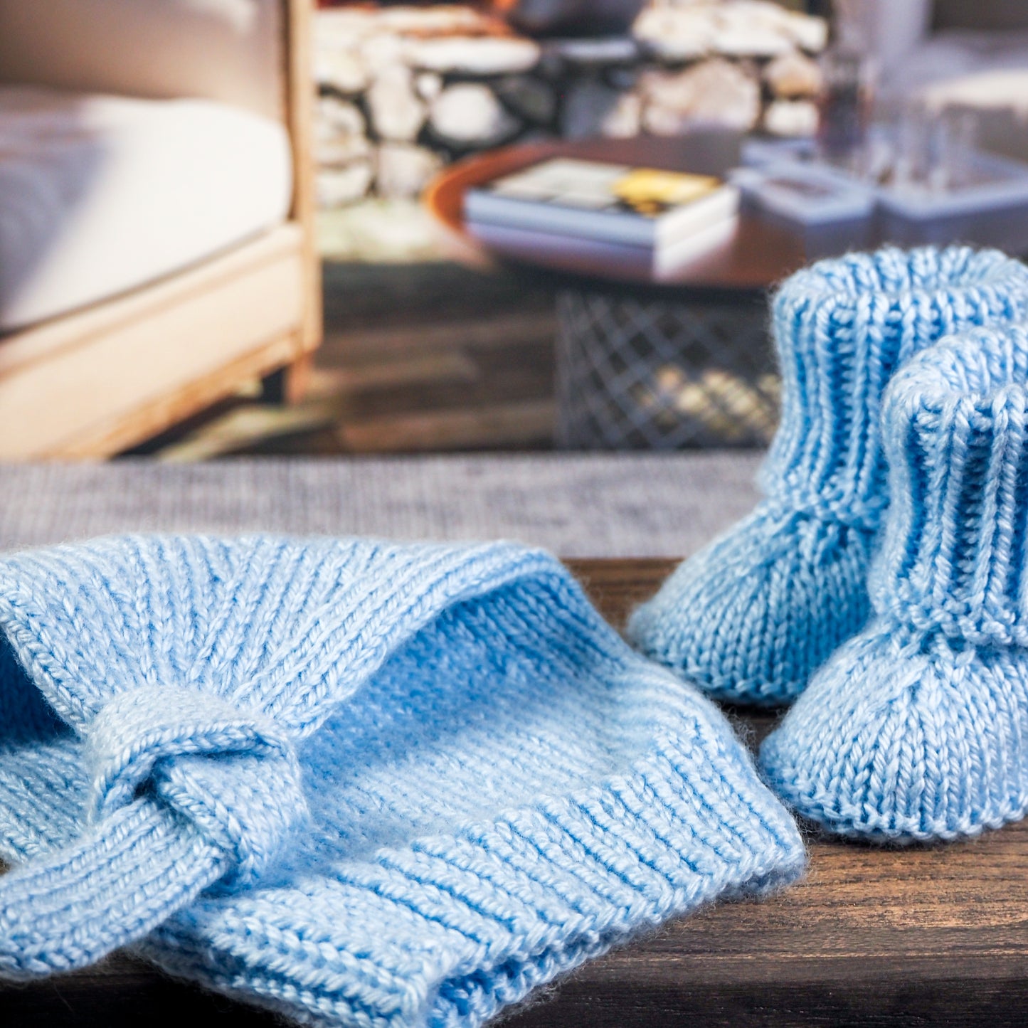 Close-up of a light blue knitted baby hat with a knotted detail and matching baby booties, displayed on a wooden surface with a cozy living room setting featuring stone walls and soft lighting in the background.