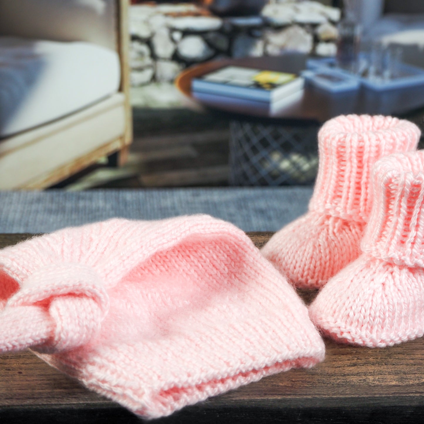 Soft pink knitted baby booties paired with a matching knotted baby hat, displayed on a wooden surface with a cozy living room background featuring a fireplace and modern decor.