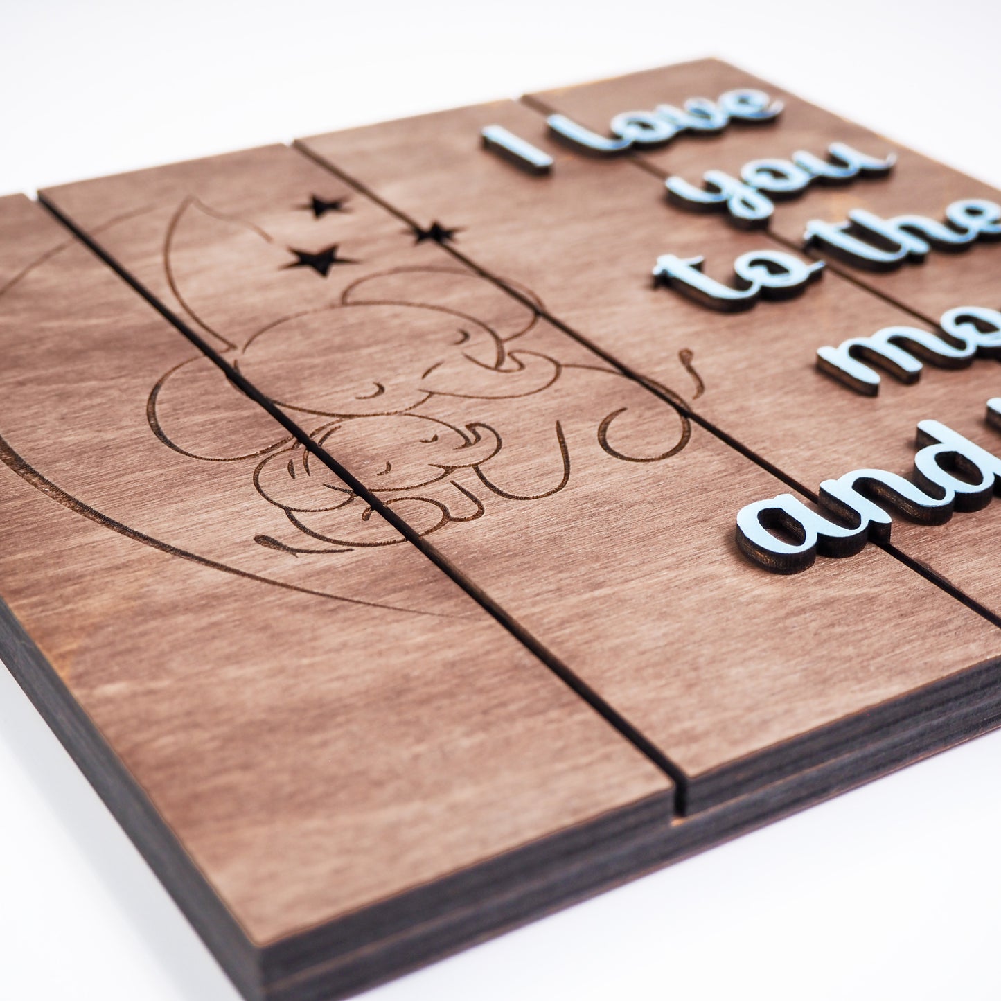 Close-up angled view of a wooden sign engraved with elephants under a crescent moon and stars, with raised blue lettering reading I love you to the moon and back.