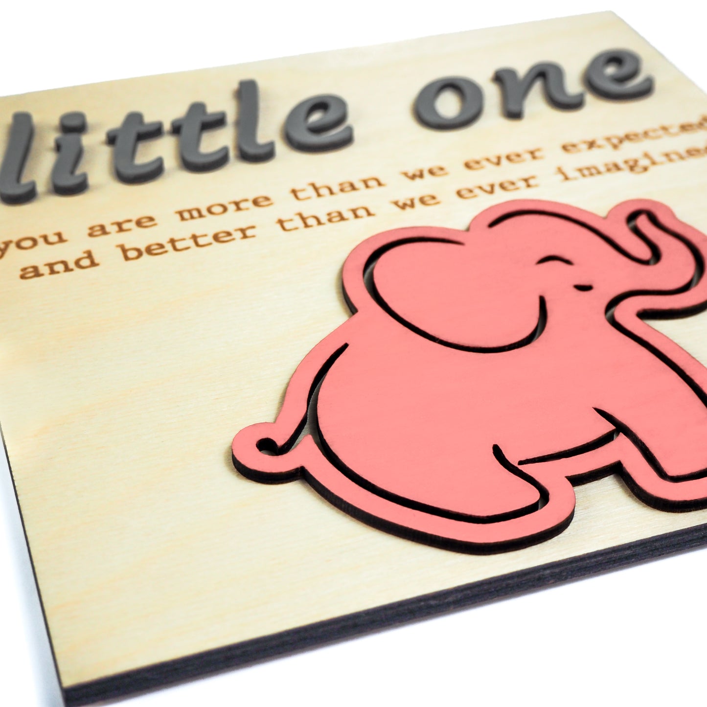 Close-up of a light wood sign with raised gray text reading "little one" and engraved text below. A pink elephant cutout is positioned near the bottom right.
