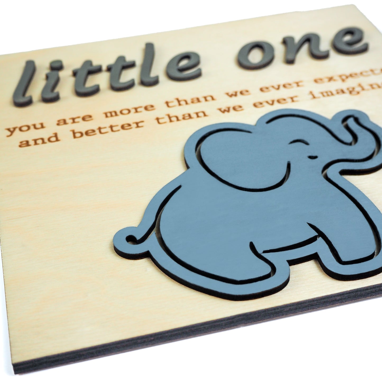 Close-up of a light wood sign with raised gray text reading "little one" and engraved text below. A blue elephant cutout is positioned near the bottom right.