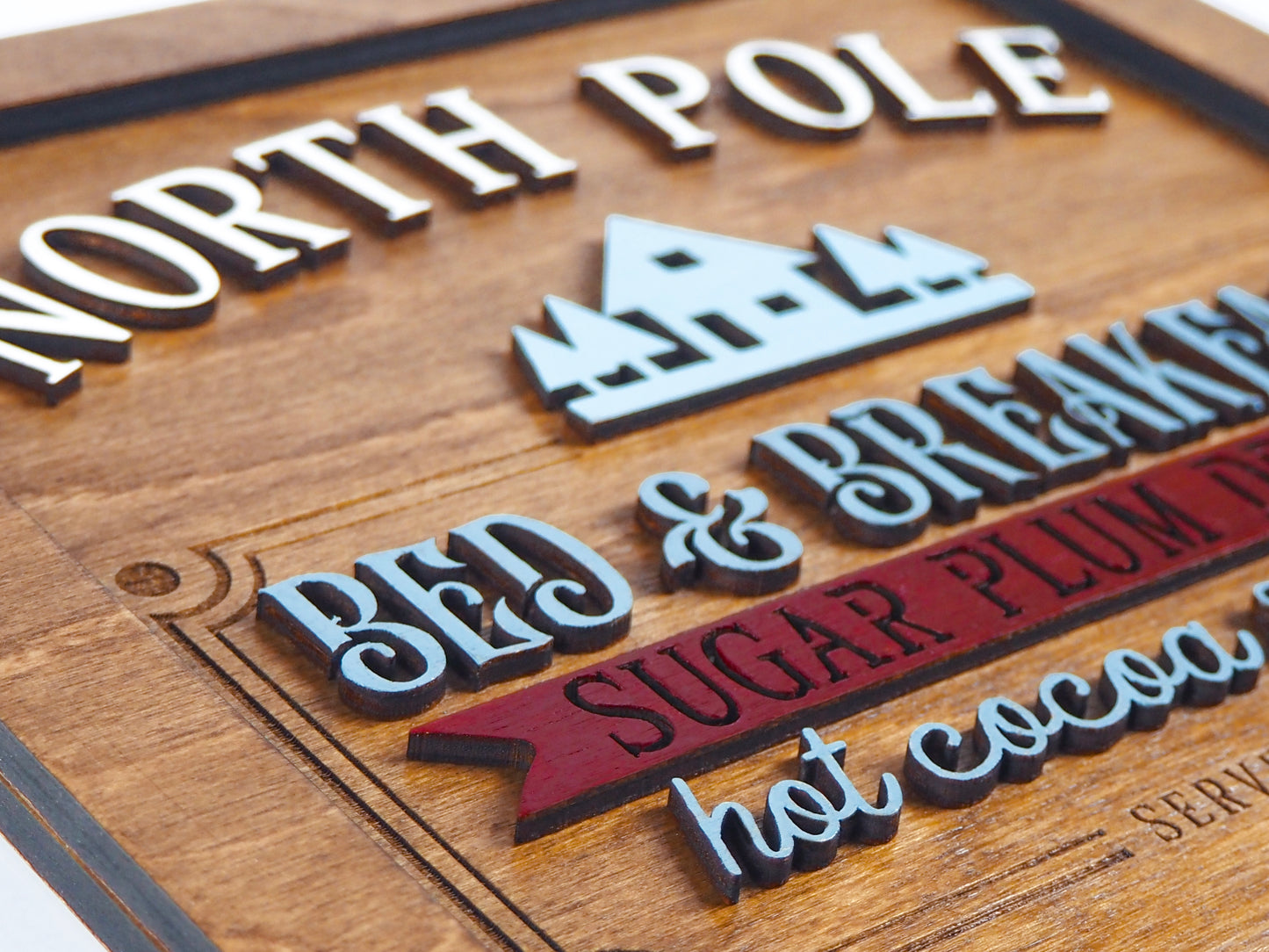 Close-up of a wooden "North Pole Bed & Breakfast" sign with raised 3D text in white, blue, and red. The words "Sugar Plum Dreams, Hot Cocoa & Cookies" are highlighted in colorful layers against the wood grain background.