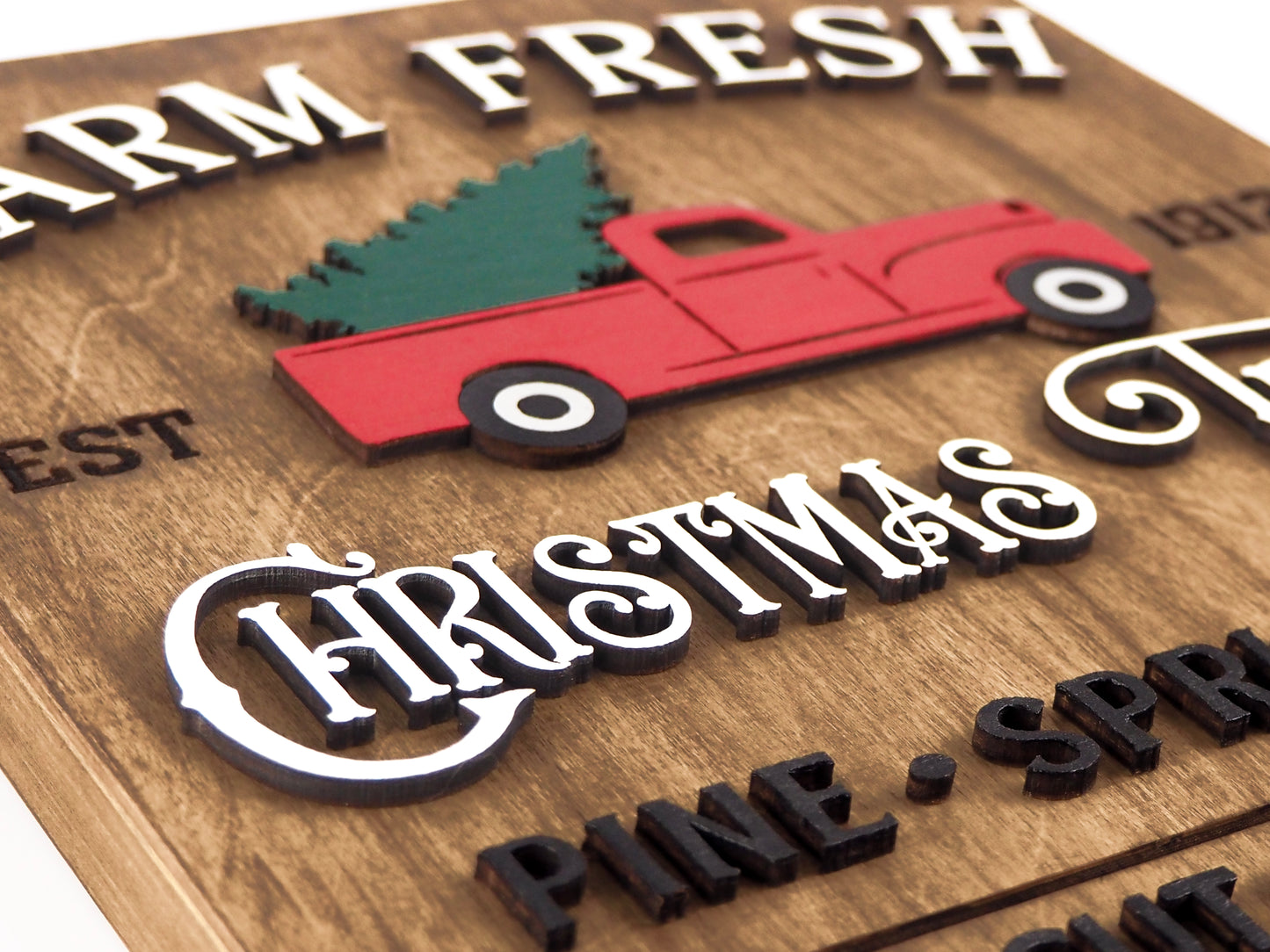 Close-up angled view of the Farm Fresh Christmas Trees sign, showcasing a raised red truck with a green tree and layered text reading "Christmas Trees." The details of the textured wooden background are visible.