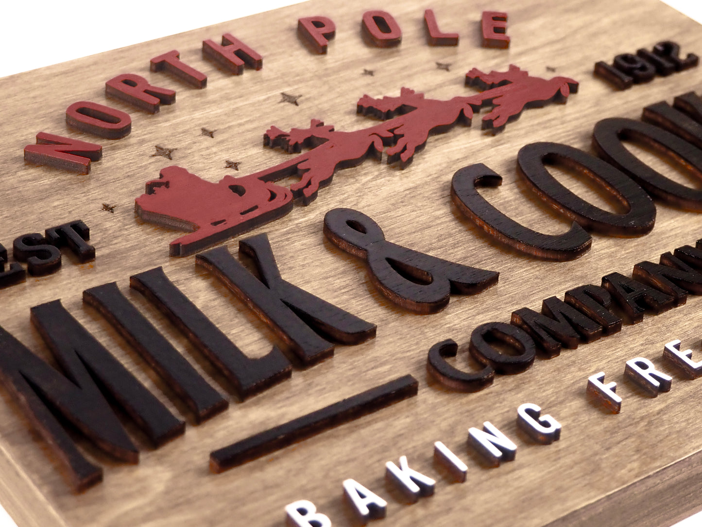Close-up angled view of the North Pole Milk & Cookie Company sign, featuring raised red Santa sleigh and reindeer with black 3D lettering on a rustic wood background. The bottom features white text reading "Baking Fresh Daily."