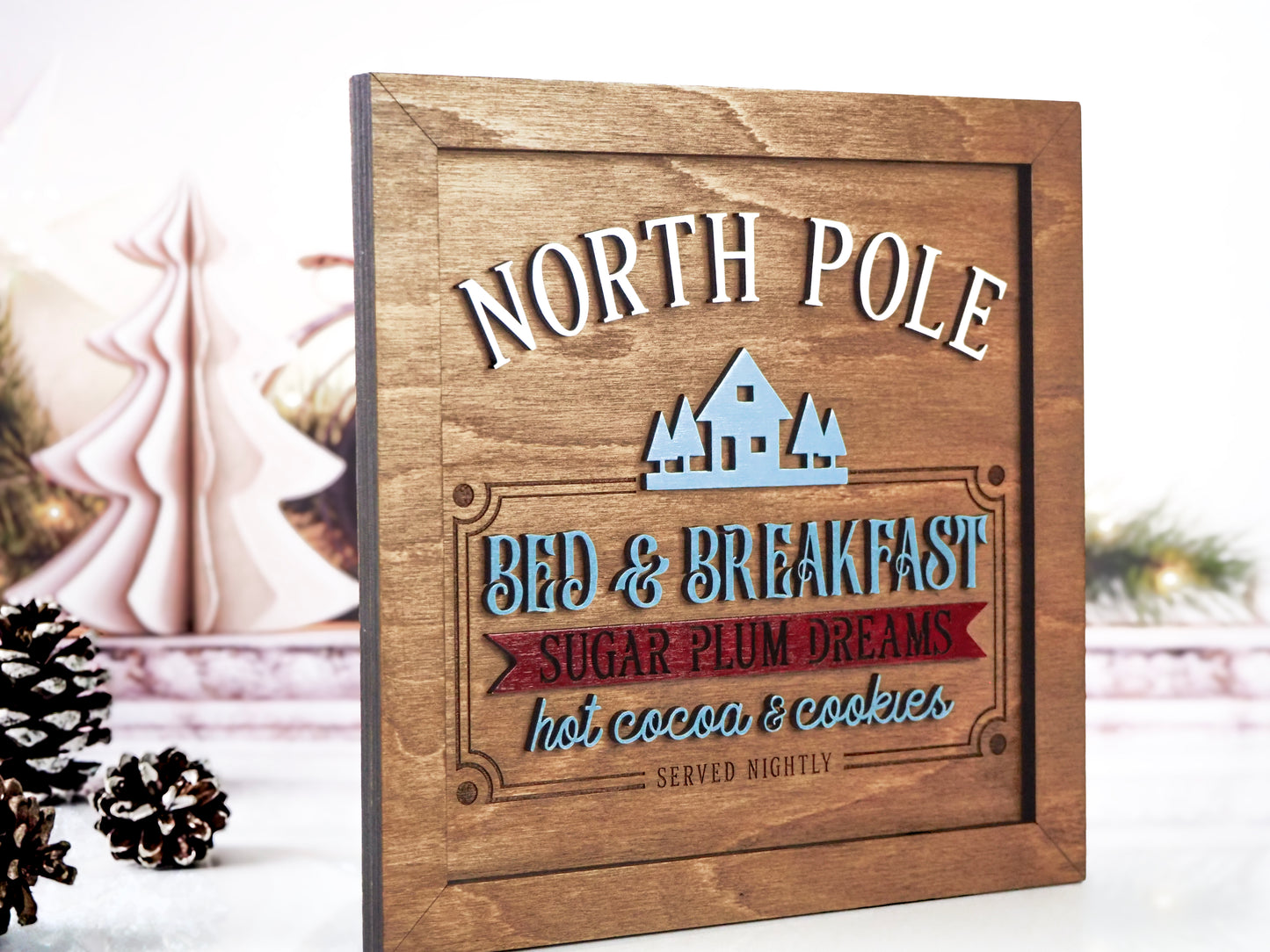 Wooden "North Pole Bed & Breakfast" sign with raised 3D text in white, blue, and red, featuring the phrases "Sugar Plum Dreams" and "Hot Cocoa & Cookies." The sign is angled against a festive holiday background with pinecones and soft Christmas décor.