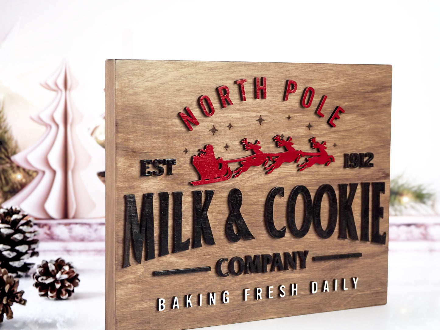 Angled view of the North Pole Milk & Cookie Company sign on a wooden background with red Santa sleigh and reindeer, black text, and white "Baking Fresh Daily" at the bottom. Decorative pinecones and holiday trees are in the background.