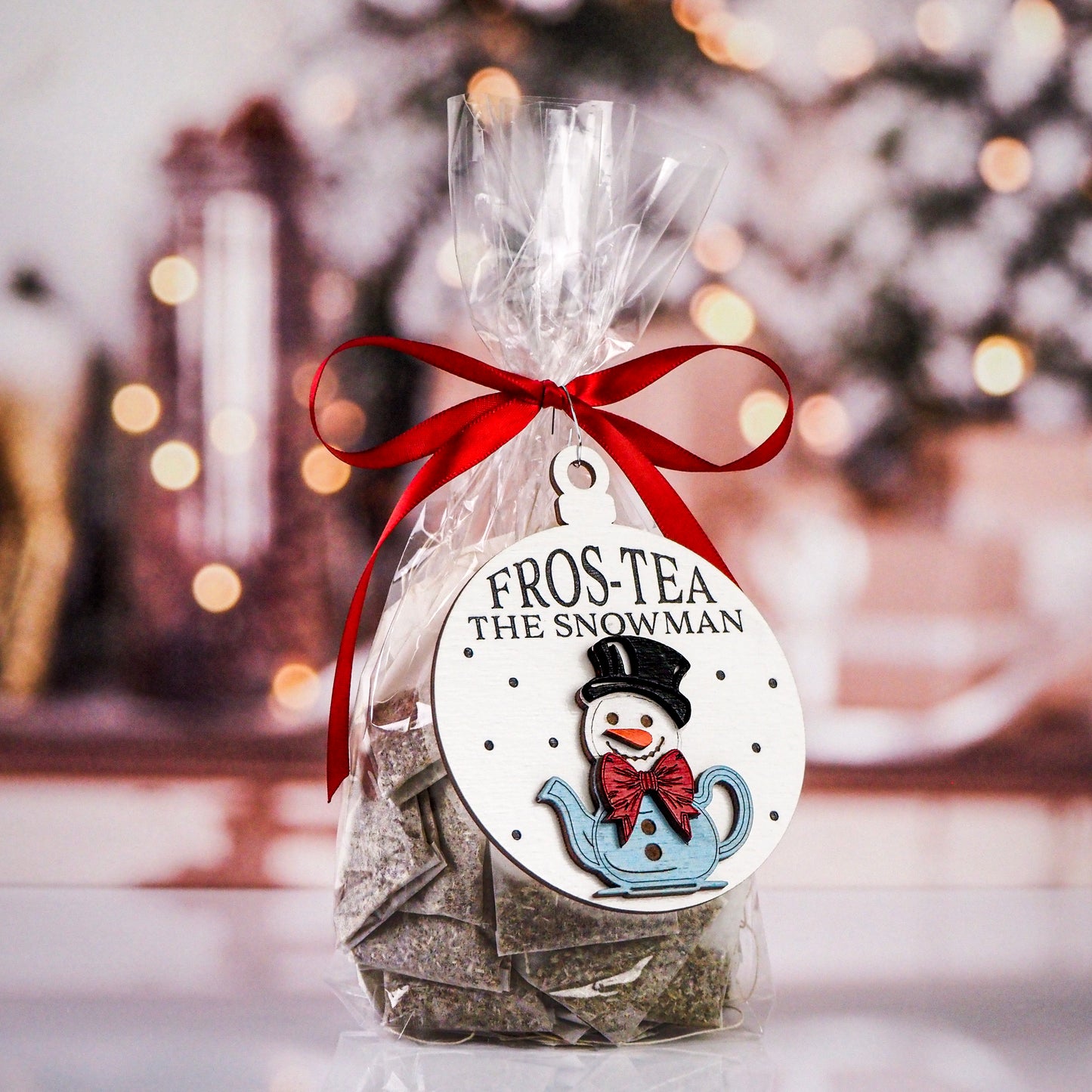 Christmas ornament featuring a snowman with a black top hat and red bow tie, sitting in a blue teapot. The ornament reads "Fros-Tea the Snowman" on a white background with dotted accents. It's attached to a gift bag of tea, tied with a red ribbon.