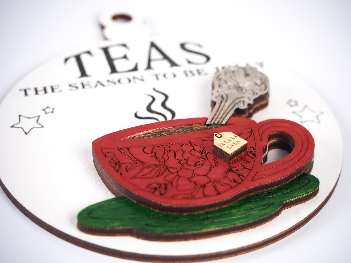 Close-up of a Christmas ornament featuring a red teacup with floral detailing, a small tea tag, and a steam design. The ornament text reads "Teas The Season To Be Jolly" in black lettering on a white background with small stars.