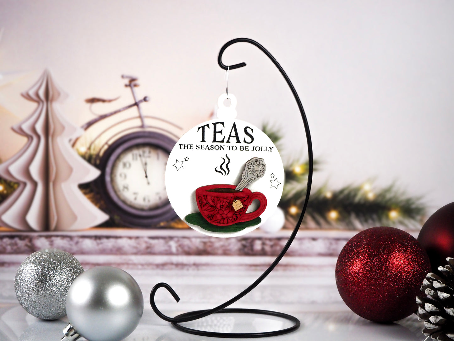 A Christmas ornament featuring a red teacup design and the words "Teas The Season To Be Jolly." The ornament hangs on a black stand with a festive background, including silver and red baubles, a pinecone, and a vintage clock.