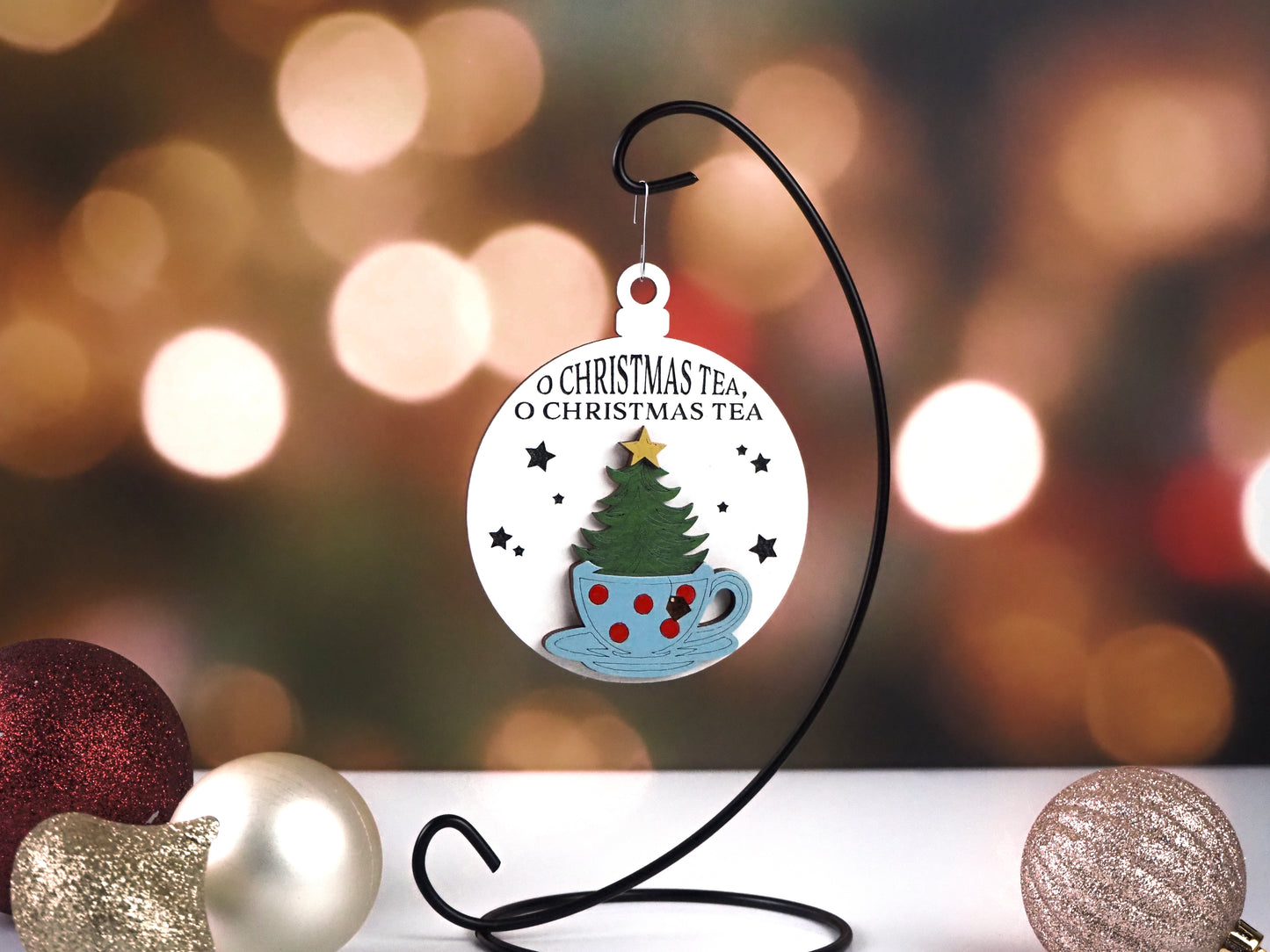 Festive Christmas ornament featuring a Christmas tree inside a polka-dotted blue teacup with a gold star topper. The ornament text reads "O Christmas Tea, O Christmas Tea" on a white background with black stars, hanging on a black metal stand with sparkling ornaments in the foreground.