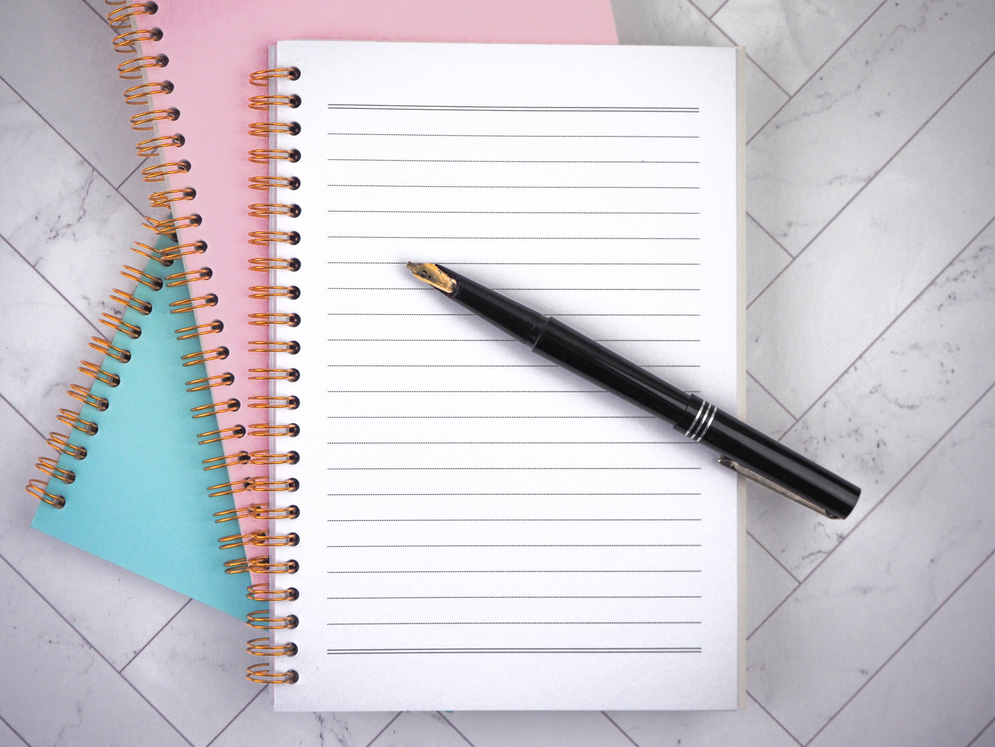 Three spiral-bound notebooks stacked together with pink, blue, and white covers. The white notebook is open, showing lined pages, with a black pen resting diagonally on it.