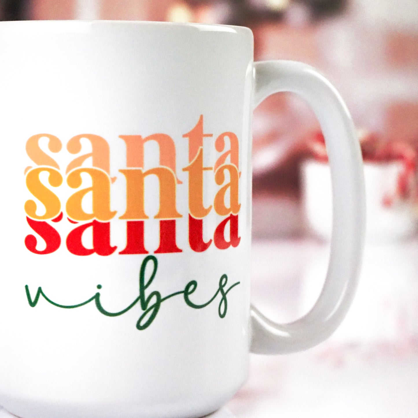 White ceramic mug with a bold "Santa Vibes" design featuring three layers of "Santa" in gradient shades of orange to red, followed by "vibes" in green cursive, set against a cozy blurred festive background.