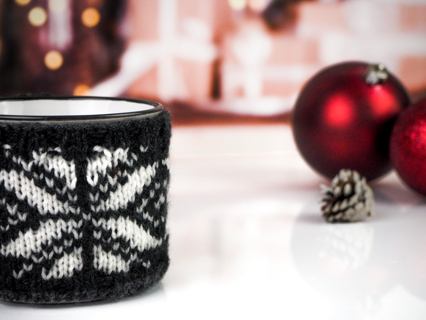 White with black speckles campfire mug wrapped in a black and white knitted cozy with a snowflake pattern, set against a festive background with red ornaments and a pine cone.
