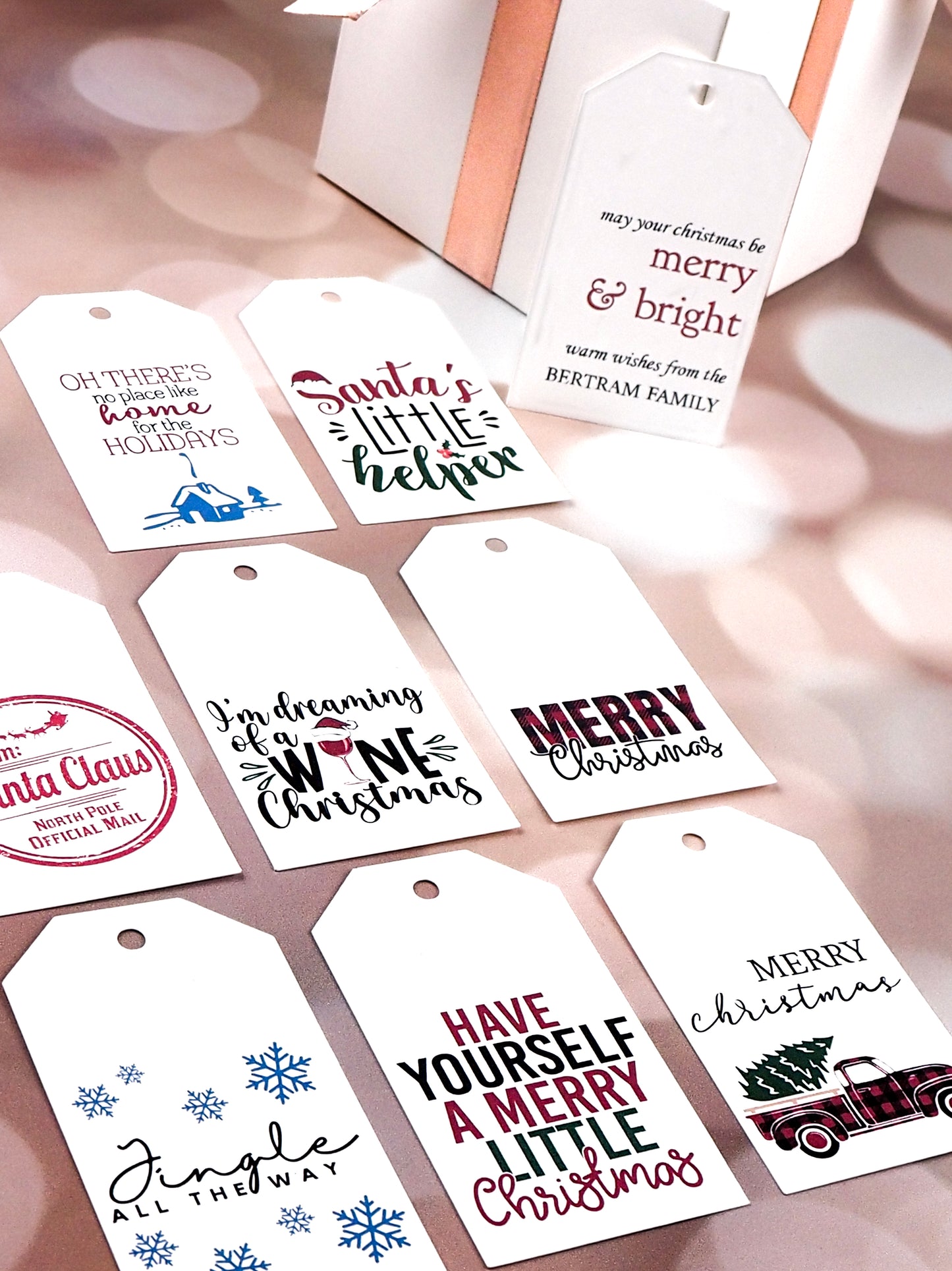 A collection of white rectangular Christmas gift tags displayed on a pink background. The designs include phrases like Oh There's No Place Like Home for the Holidays with a house graphic, Santa's Little Helper with a Santa hat, Merry & Bright with warm wishes from the Bertram Family, I'm Dreaming of a Wine Christmas with a wine glass graphic, Merry Christmas, Jingle All The Way with blue snowflakes, Have Yourself a Merry Little Christmas, and Merry Christmas with a red truck carrying a Christmas tree.