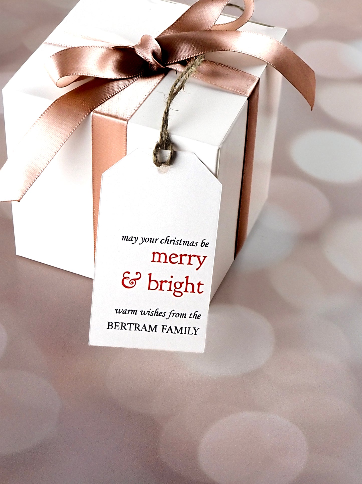 White gift box tied with a pink satin ribbon and adorned with a white rectangular gift tag. The tag reads, "May your Christmas be Merry & Bright. Warm wishes from the Bertram Family" in red and black text.