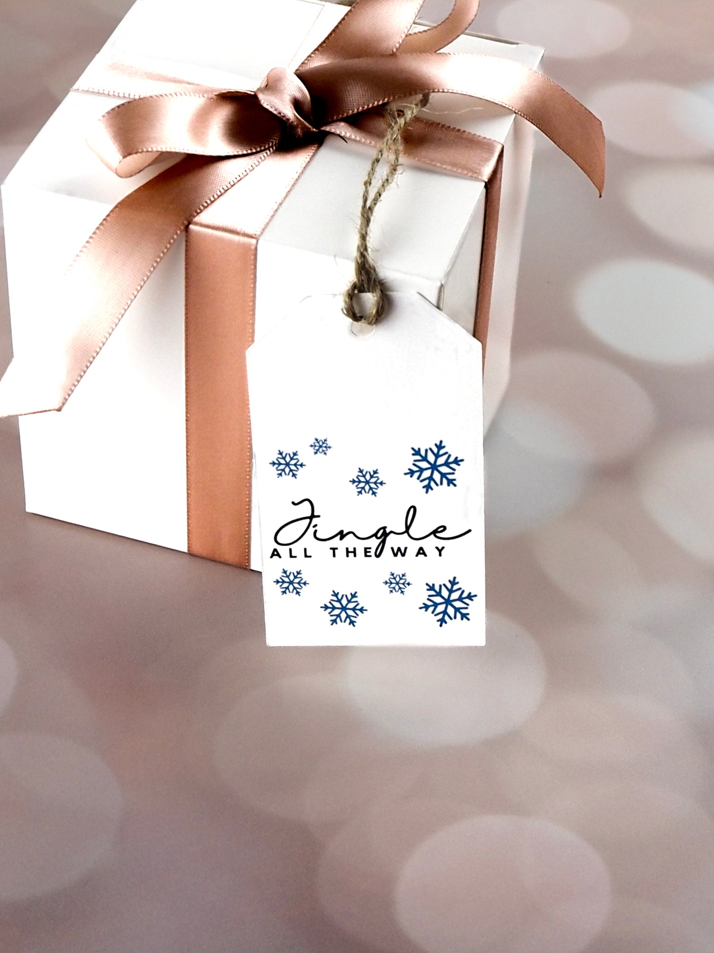 White gift box tied with a pink satin ribbon and adorned with a white rectangular gift tag. The tag features the phrase "Jingle All The Way" in black script, surrounded by blue snowflakes.