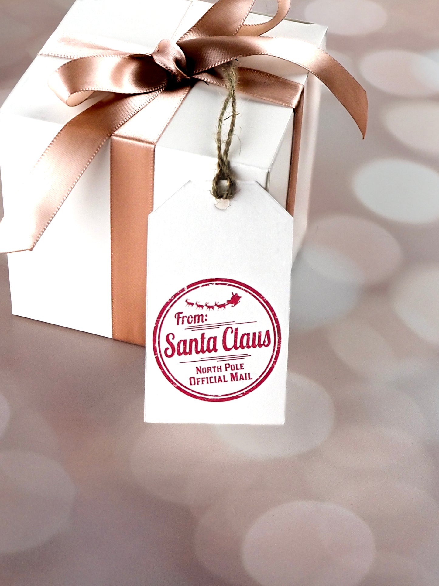 White gift box tied with a pink satin ribbon and adorned with a white rectangular gift tag. The tag features a red circular stamp design that reads "From Santa Claus, North Pole Official Mail" with a silhouette of Santa's sleigh and reindeer.