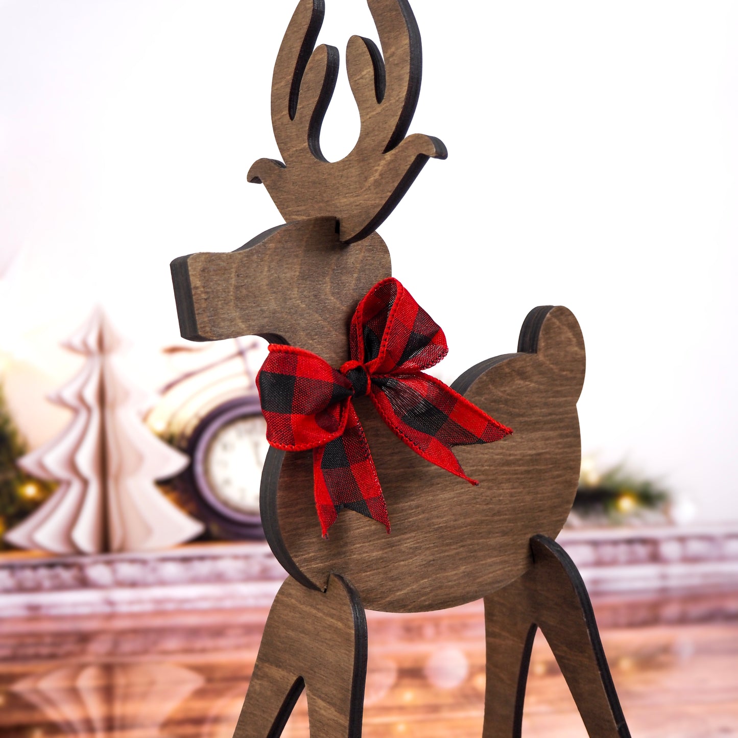Close-up of a wooden reindeer with a red and black buffalo plaid bow, set against a festive background featuring Christmas trees and holiday décor.