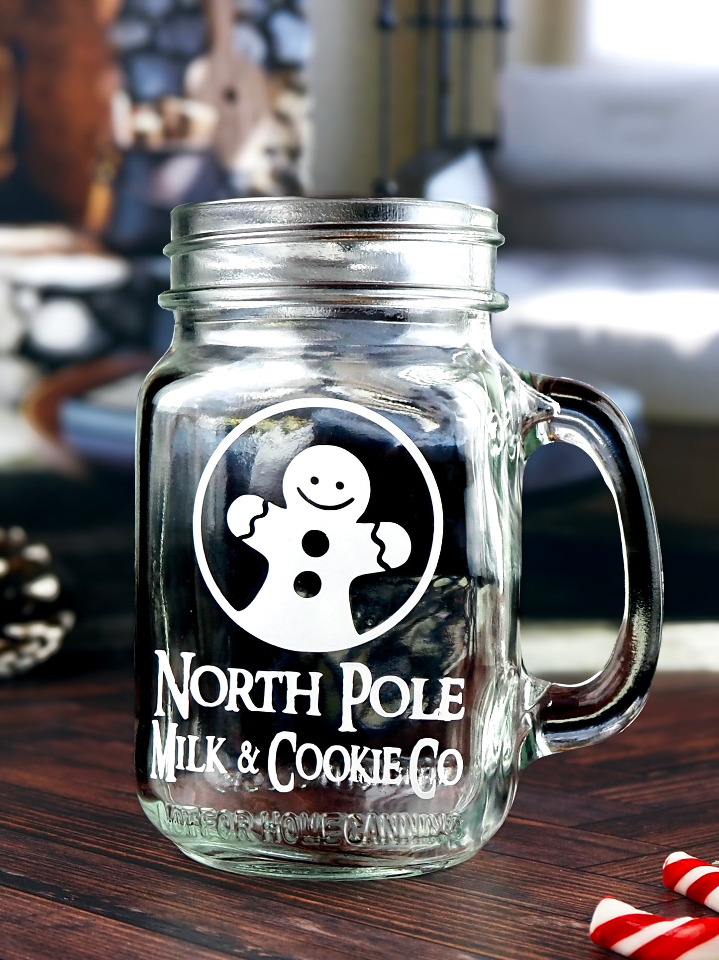 Clear glass mason jar-style mug with a handle. The mug features a white graphic of a smiling gingerbread man inside a circle, along with the text North Pole Milk & Cookie Co in a vintage-style font.