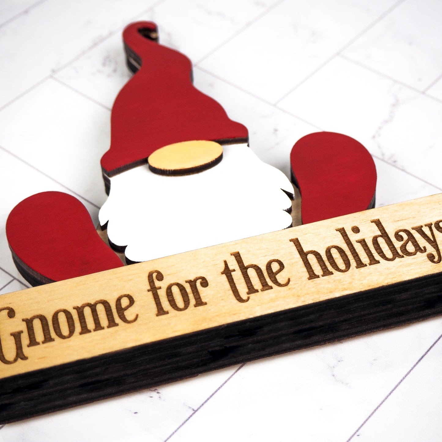 Close-up of a festive gnome tabletop decoration featuring a red hat, white beard, and a wooden base engraved with Gnome for the holidays. The background showcases a tiled surface, emphasizing the detailed craftsmanship of the gnome and its engraving.