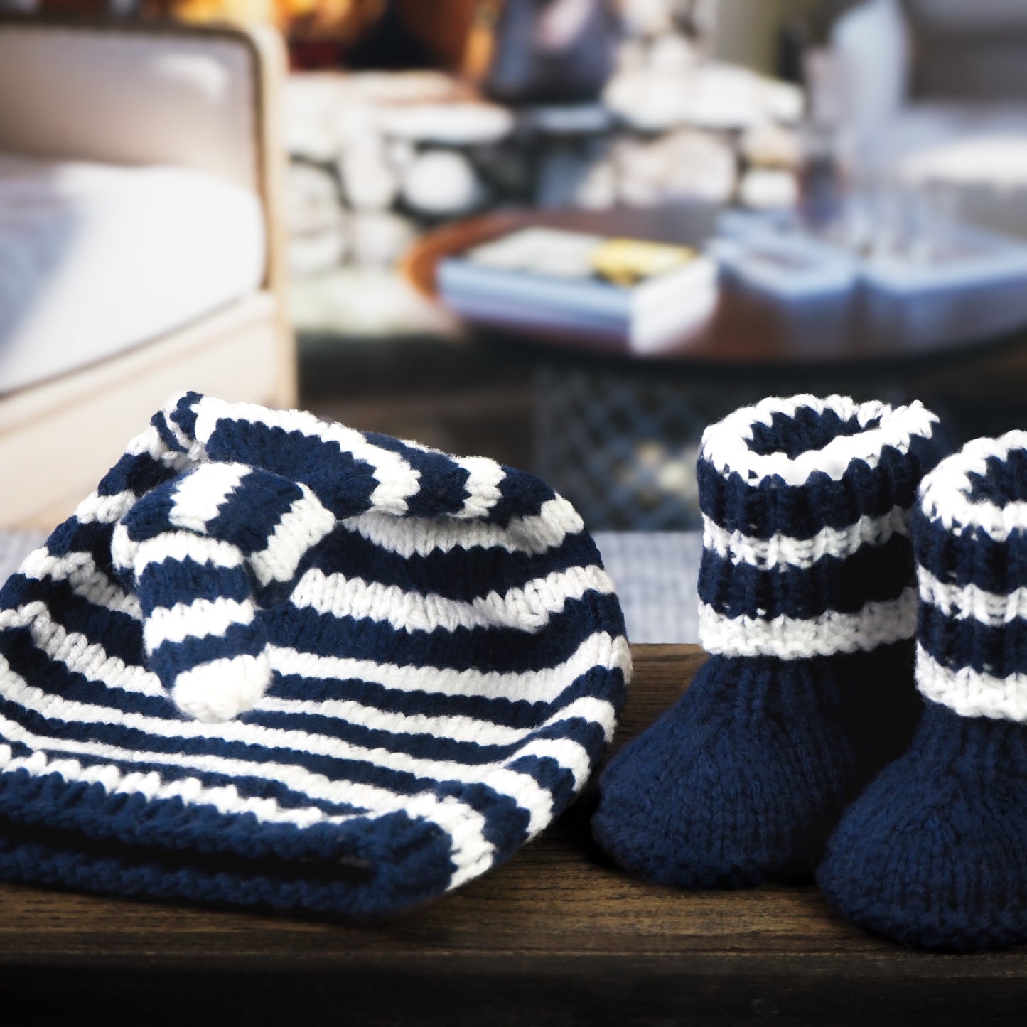 A cozy navy blue and white striped hand-knitted baby hat with a knot and matching booties, displayed on a wooden surface with a warm living room and fireplace in the background.