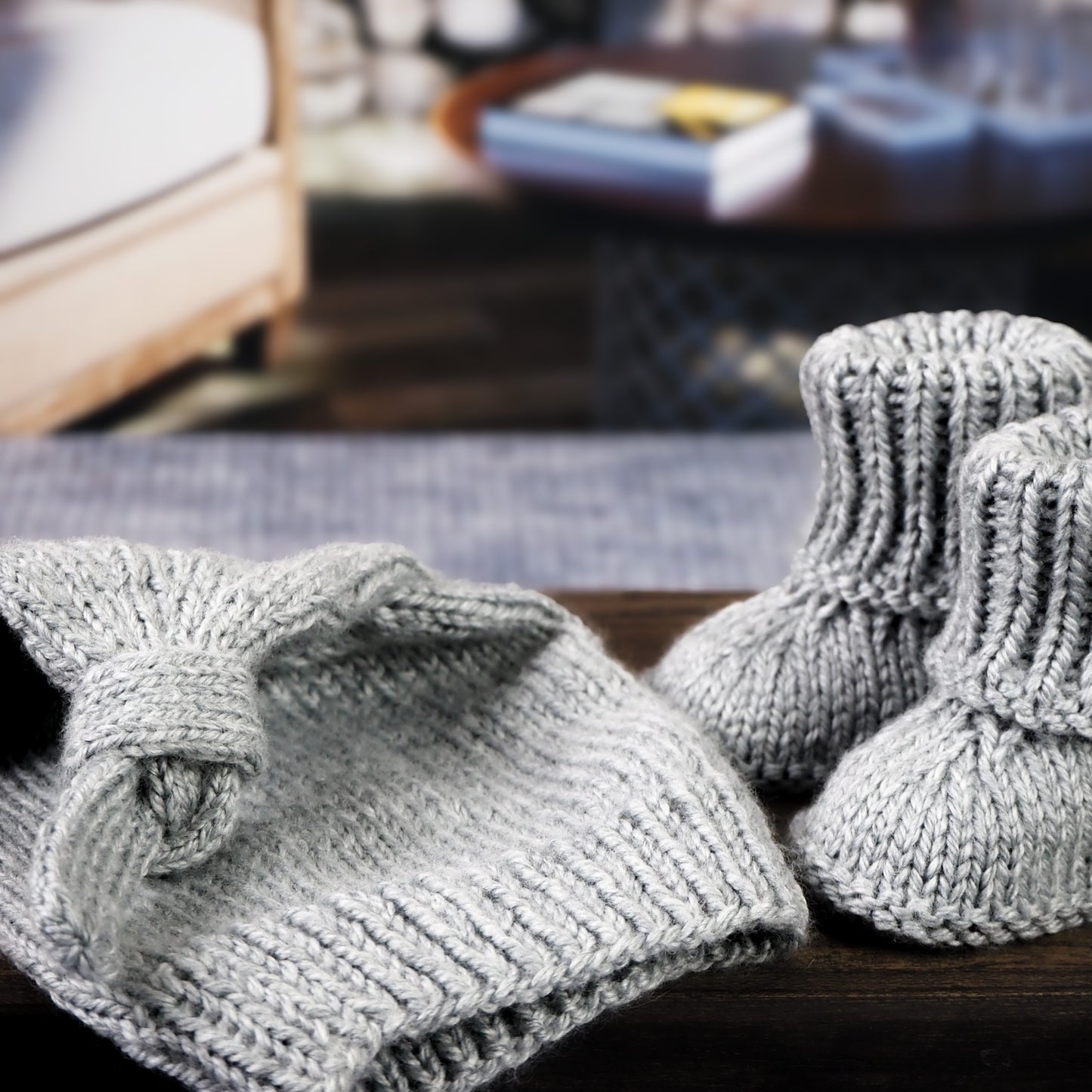 Close-up of a gray knitted baby hat with a knot detail and matching booties, displayed on a wooden surface in front of a cozy living room setting with warm tones and soft lighting.