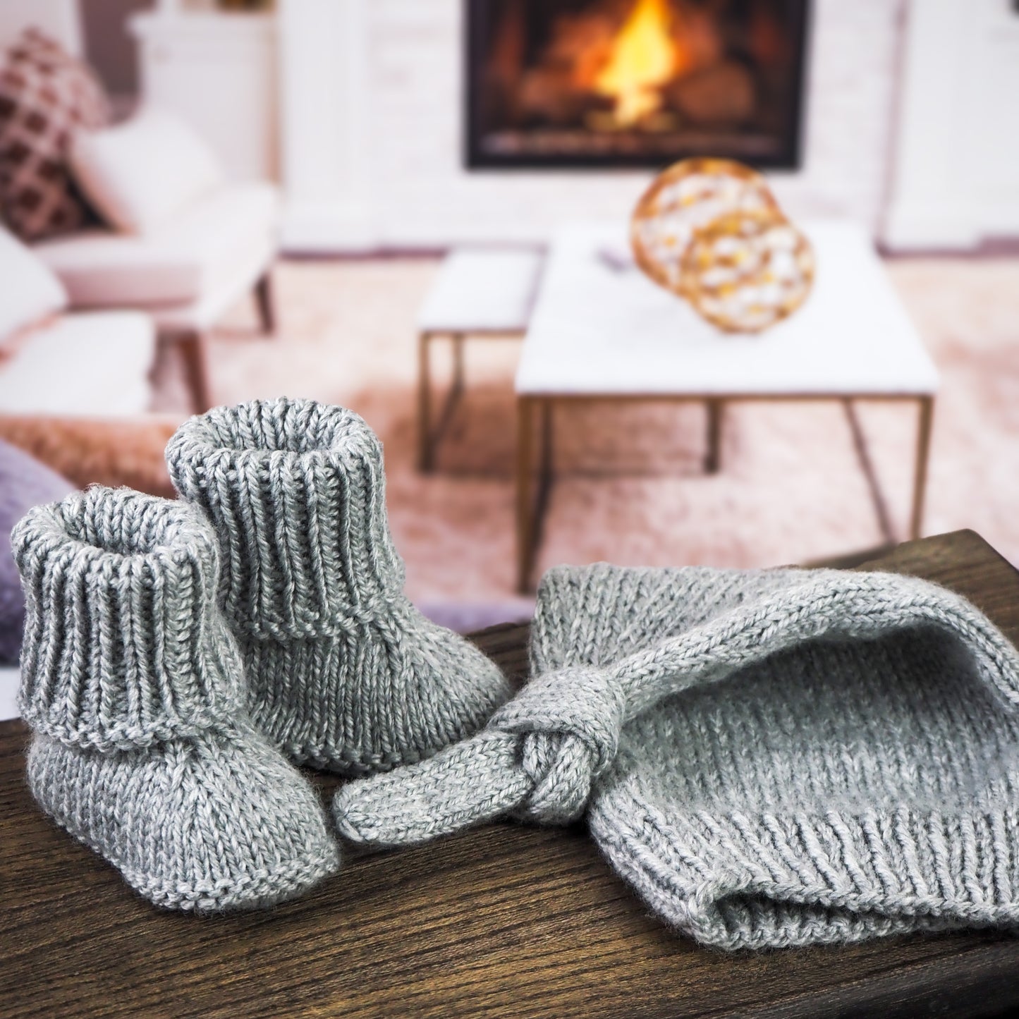 Gray knitted baby hat with a knot detail and matching booties displayed on a wooden surface in front of a cozy living room setting with a lit fireplace in the background.
