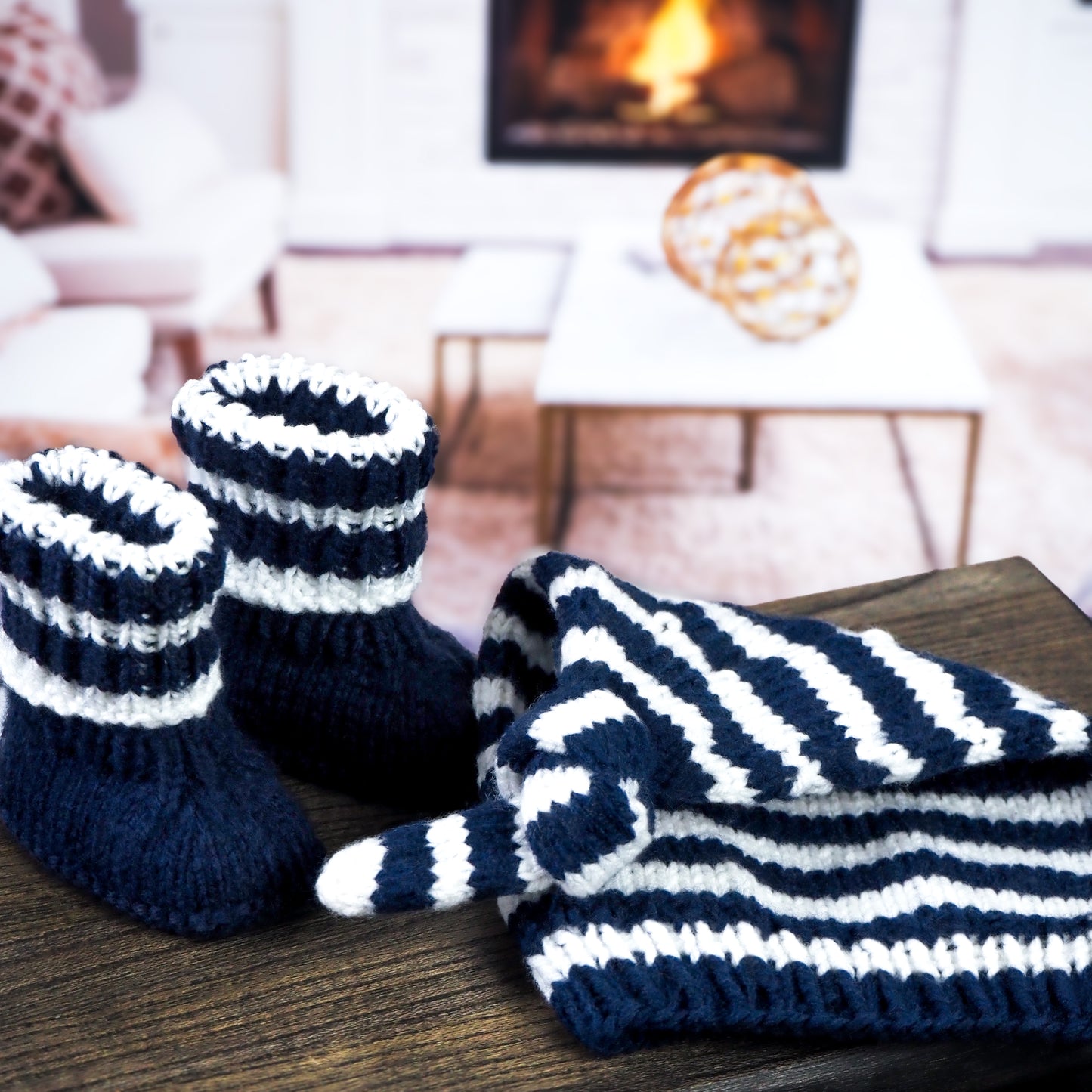 A close-up of a gray hand-knitted baby hat with a knot and matching booties, displayed on a reflective black surface with a blurred cozy indoor background.