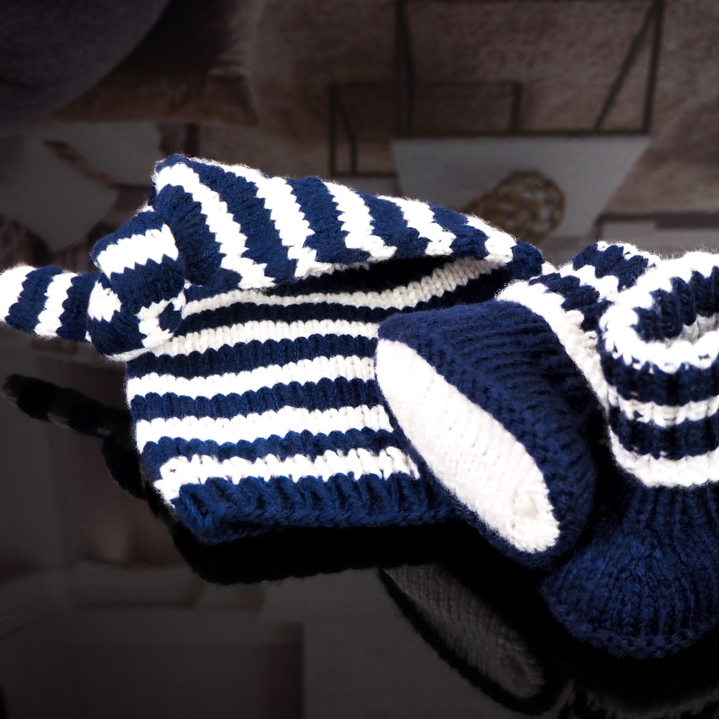 A navy blue and white striped hand-knitted baby hat with a knot and matching booties, neatly displayed on a wooden surface with a blurred cozy living room and coffee table in the background.