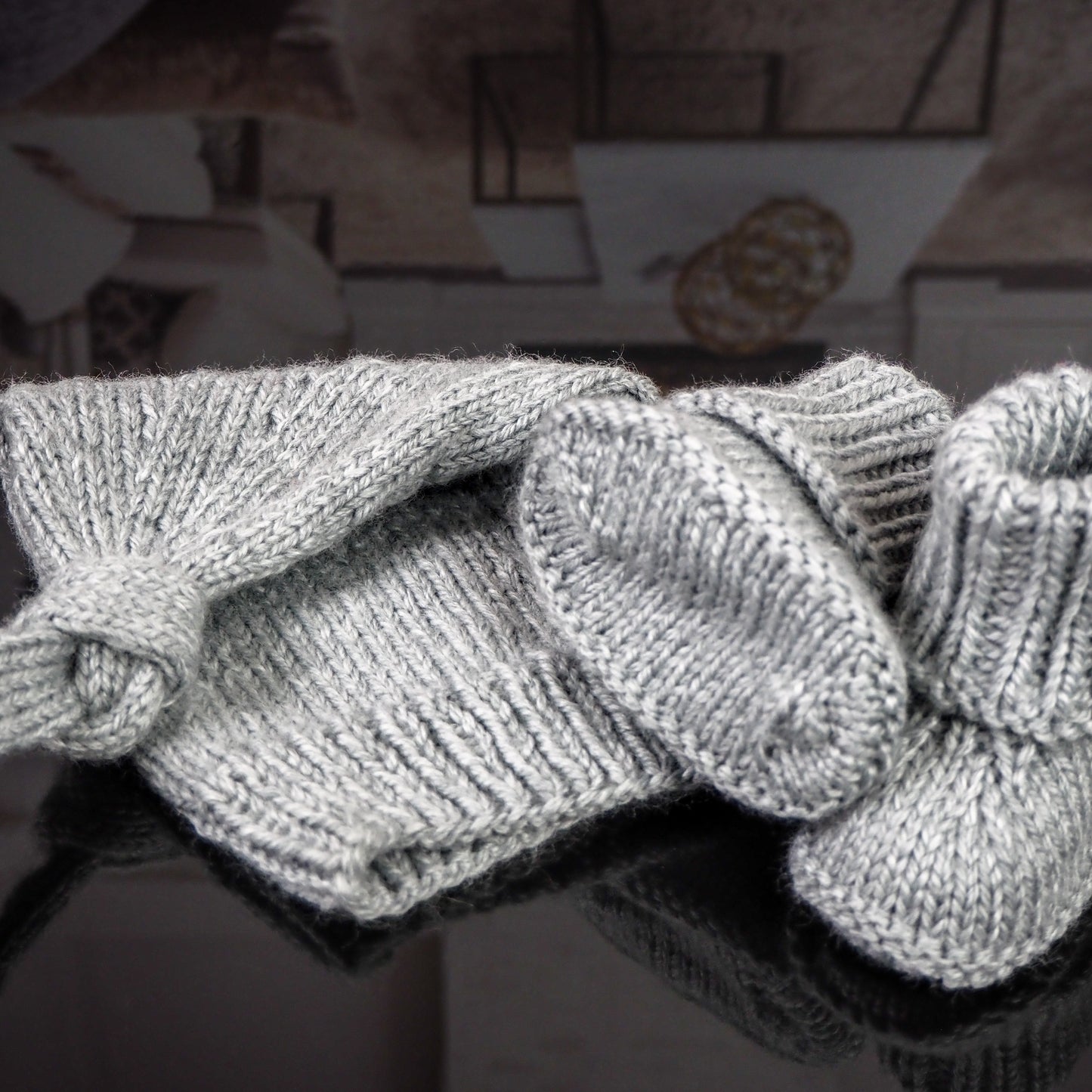 Gray knitted baby hat with a knot detail and matching booties, displayed on a reflective black surface with a cozy living room background in warm tones.