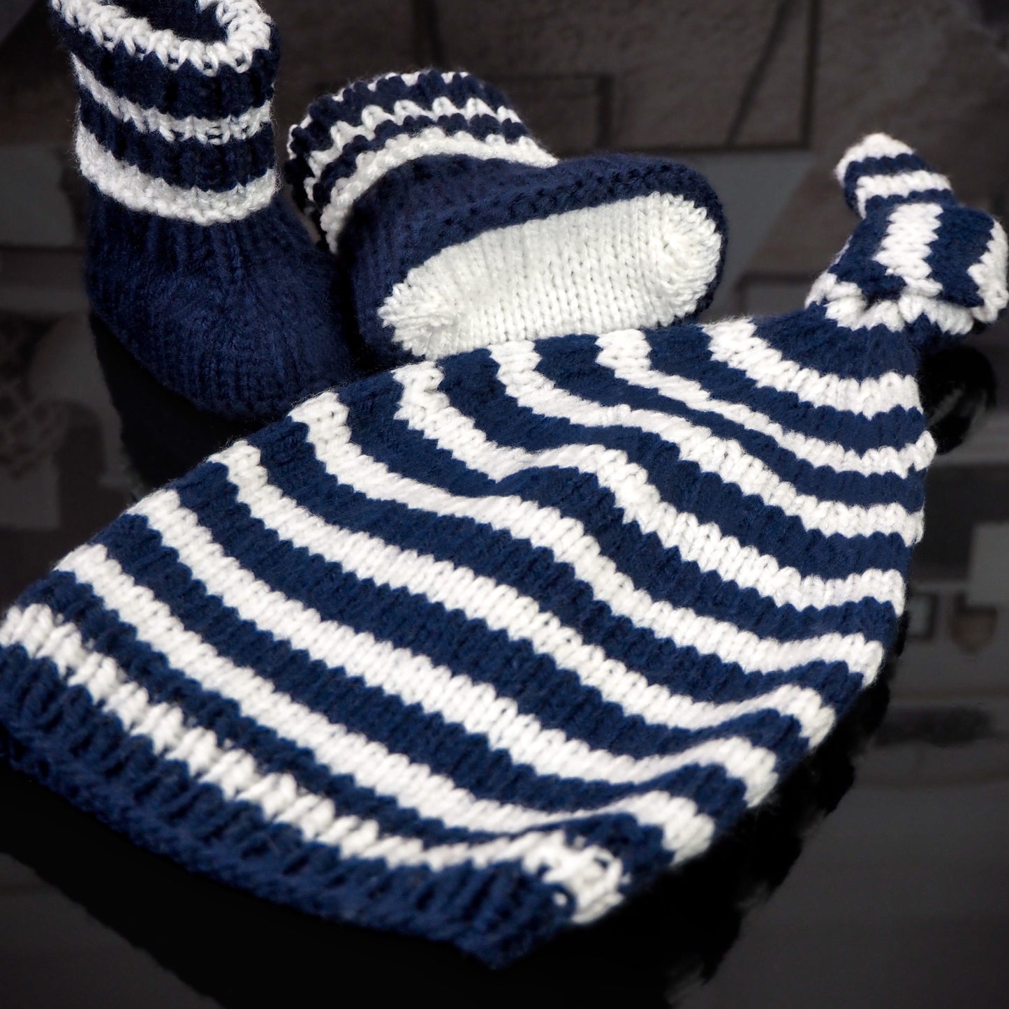 Close-up of a navy blue and white striped hand-knitted baby hat with a knot and matching booties, placed on a glossy black surface with a blurred background featuring cozy interior decor.