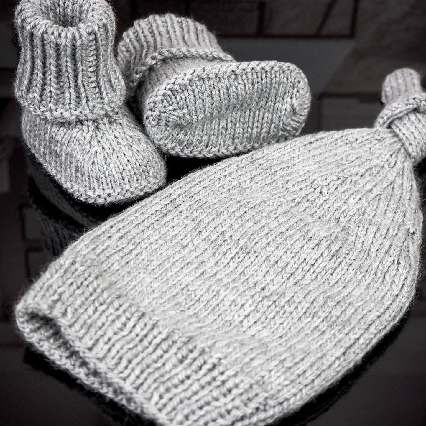 Gray knitted baby hat with a knot detail and matching booties, displayed on a reflective surface with a blurred background, highlighting the soft texture and craftsmanship.