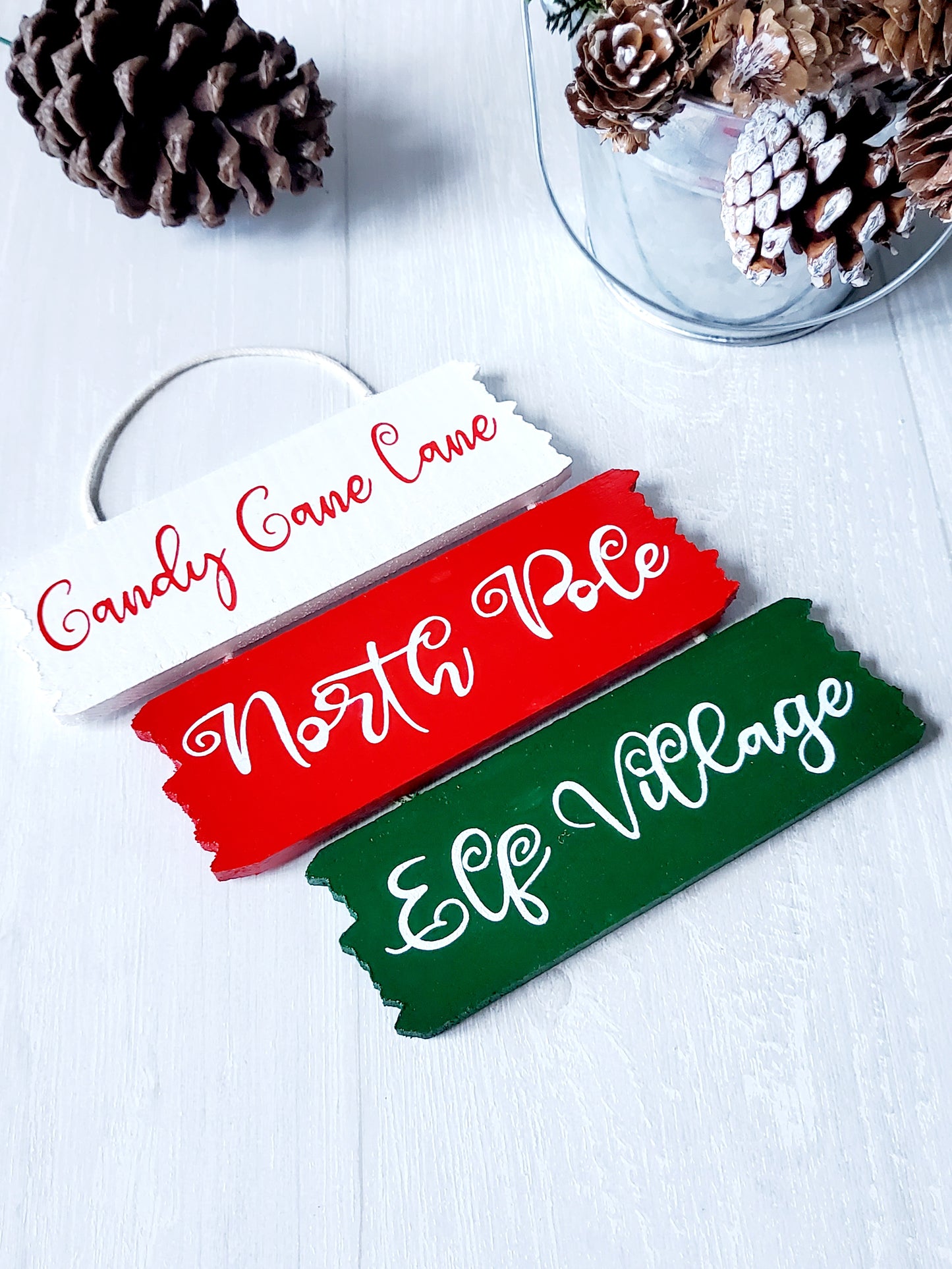 3 wooden hanging décor signs painted each in white, red and green are 2-inches in height and 6.25-inches in length. Displaying the phrases Candy Cane Lane, North Pole and Elf Village in white. Hand-painted by Snuggle Up Co.