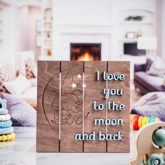 Wooden nursery sign with engraved elephants cuddling under a crescent moon and stars, and the phrase I love you to the moon and back in light blue text, displayed on a marble surface with cozy toys and a fireplace in the background.