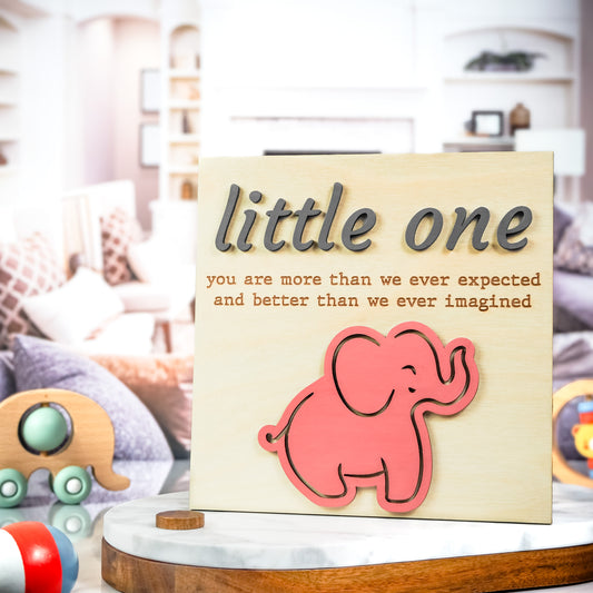 Wooden sign with raised gray and engraved lettering that reads "little one you are more than we ever expected and better than we ever imagined," featuring a pink elephant illustration at the bottom.