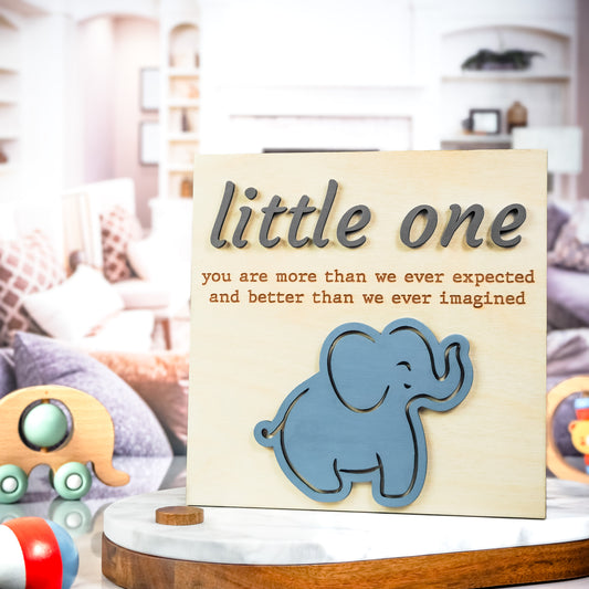 Wooden sign with raised gray and engraved lettering that reads "little one you are more than we ever expected and better than we ever imagined," featuring a blue elephant illustration at the bottom.
