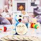 Blue stuffed bear holding a wooden milestone disc engraved with one month and a baby elephant illustration, surrounded by additional milestone discs and toys in a cozy living room setting.