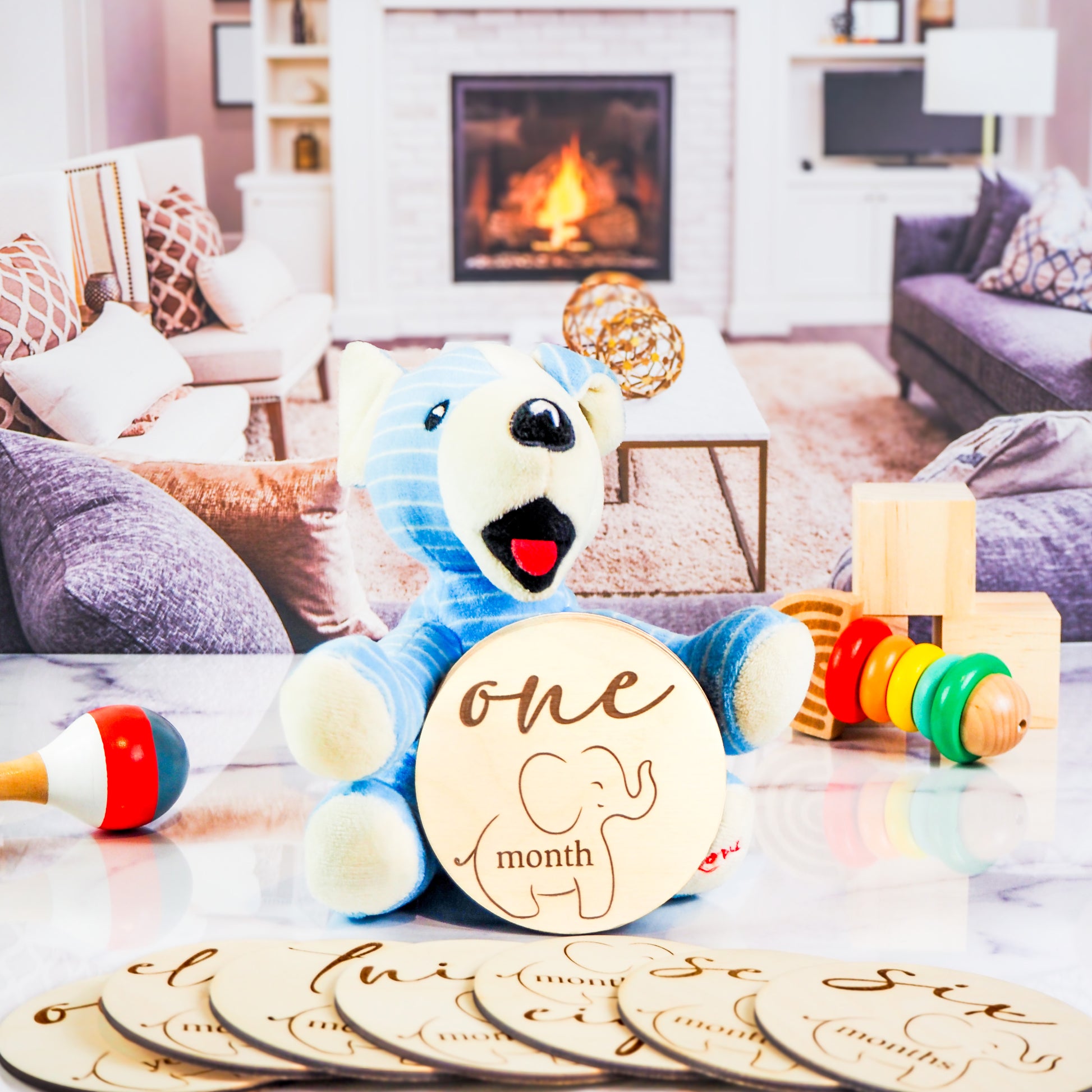 Blue stuffed bear holding a wooden milestone disc engraved with one month and a baby elephant illustration, surrounded by additional milestone discs and toys in a cozy living room setting.