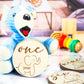 Blue stuffed bear holding a wooden milestone disc engraved with "one year" and a baby elephant design, surrounded by other milestone discs and toys.