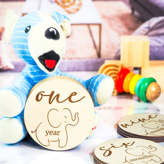 Blue stuffed bear holding a wooden milestone disc engraved with "one year" and a baby elephant design, surrounded by other milestone discs and toys.