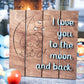 Wooden sign featuring engraved elephants cuddling under a crescent moon and stars, with the phrase I love you to the moon and back in blue lettering, displayed on a soft blue rug with baby toys and a cozy fireplace in the background.
