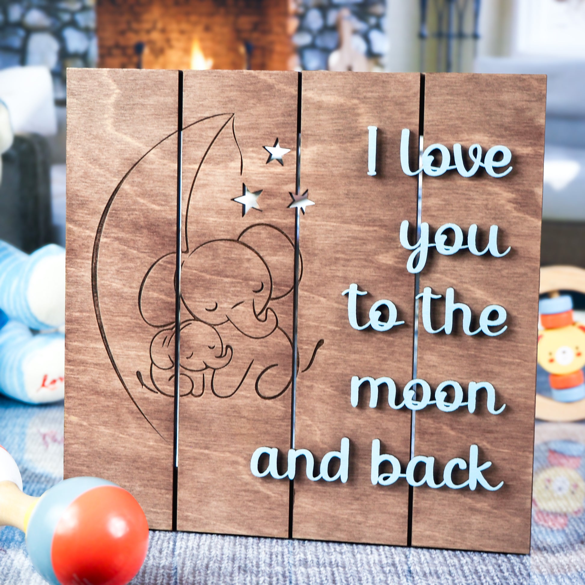 Wooden sign featuring engraved elephants cuddling under a crescent moon and stars, with the phrase I love you to the moon and back in blue lettering, displayed on a soft blue rug with baby toys and a cozy fireplace in the background.