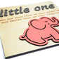 Close-up of a light wood sign with raised gray text reading "little one" and engraved text below. A pink elephant cutout is positioned near the bottom right.