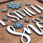 Close-up view of the "I Smell Snow" sign featuring raised white letters and blue snowflakes on a wooden background. The details of the layered design are highlighted.