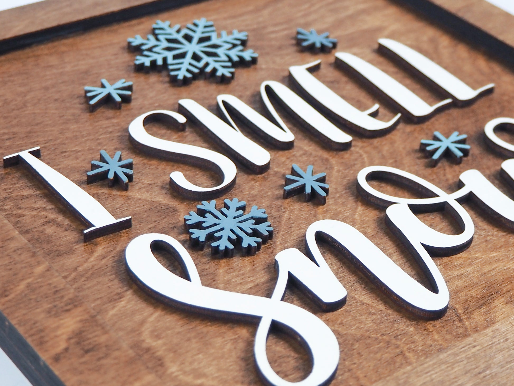 Close-up view of the "I Smell Snow" sign featuring raised white letters and blue snowflakes on a wooden background. The details of the layered design are highlighted.